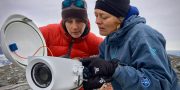 Two women in red and blue jackets with woolly hats examine a big grey camera