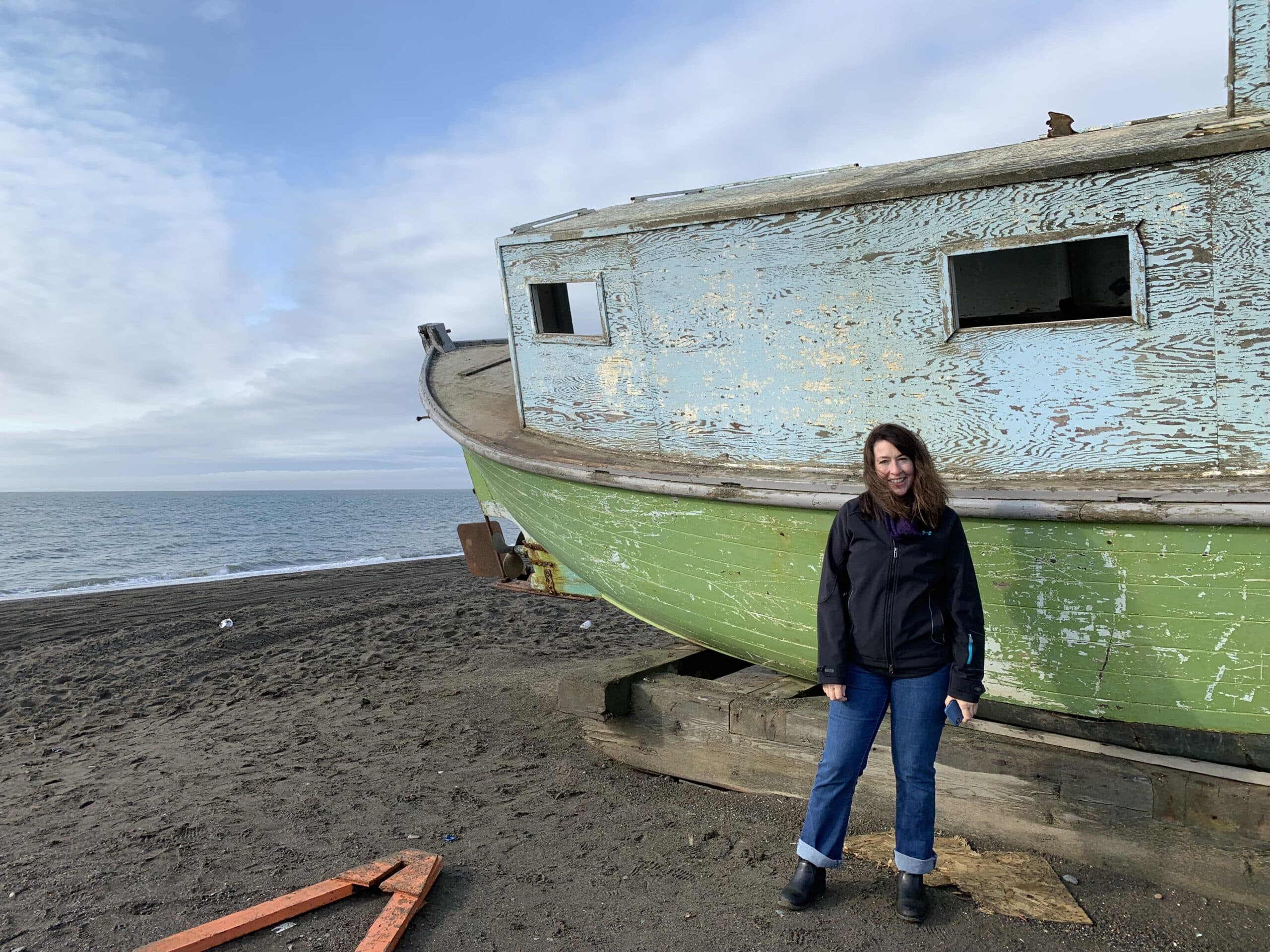 A woman with dark hair in a dark jacket and blue jeans stands in front of a blue and green fishing boat in front of a blue sky
