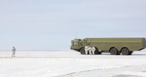 Four soldiers in white uniforms stand by a large green military truck on white snowy ground against a pale blue background