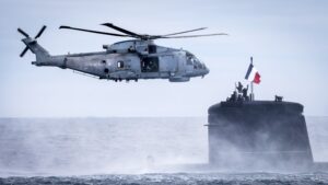 A gray British Royal Navy Merlin MK2 Helicopter hovering next to the Amethyste Attack Submarine of the French Navy, during the NATO Steadfast Defender 24 exercise off the coast of Norway in 2024 over a dark blue sea