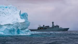 Grey ship behind blue-white iceberg against grey sky in waters around Greenland