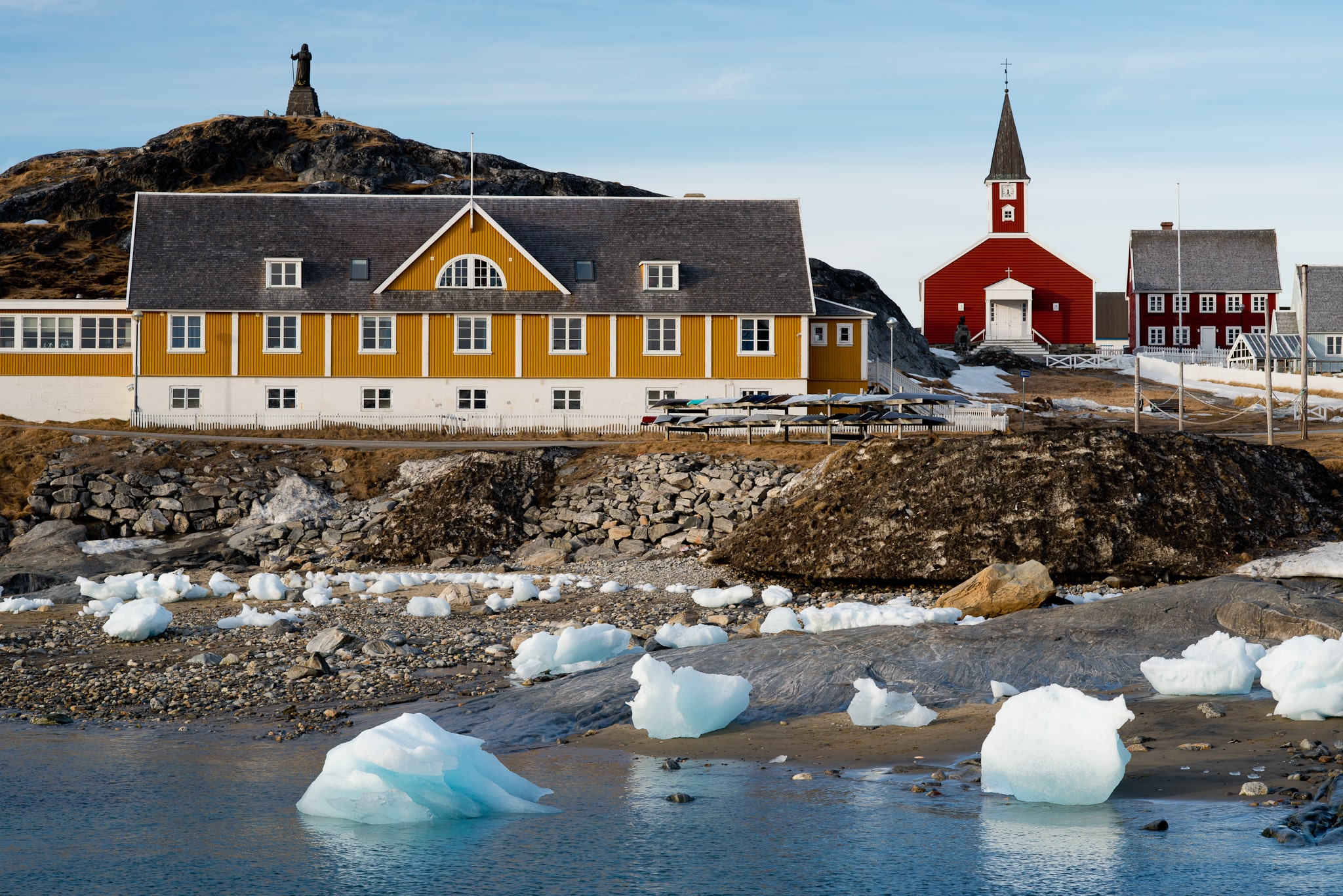 Orange and red buildings on land in front of small glaciers in water in the foreground with a statue in the background