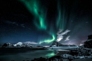 Stunning view of the Northern Lights over snow-capped mountains and calm waters, with green auroras dancing across a starry Arctic sky, reflecting on the landscape